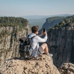 A man sits on a cliff edge taking a photo of a scenic canyon with his phone, capturing a breathtaking landscape.