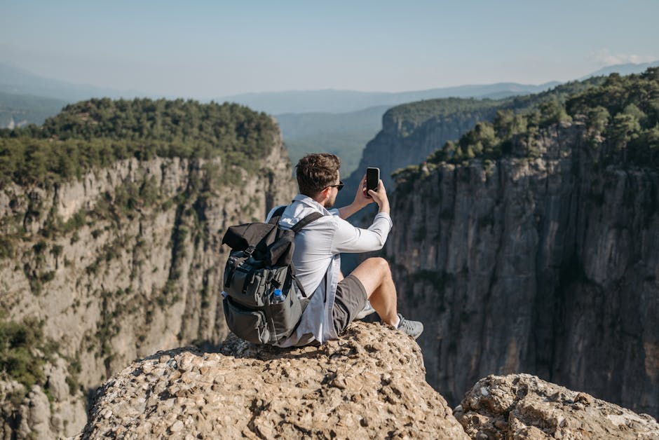 A man sits on a cliff edge taking a photo of a scenic canyon with his phone, capturing a breathtaking landscape.