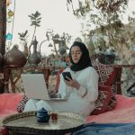 The Nomad’s Trap: Why Remote Work is the Ultimate Architectural Psychological Challenge A woman in a hijab working on a laptop and phone in a cozy outdoor setting.