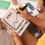 The Personalization Paradox: Scaling High-Performance Systems Close-up of a man drawing a marketing strategy graph in a notebook.