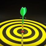 Close-up of a green dart hitting the bullseye on a yellow and black target board, symbolizing precision and success.