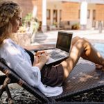 The Productivity Paradox: Why Leisure Drives High Performance Woman with curly hair working on a laptop outdoors by the poolside, embracing remote work lifestyle.
