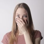 Surprised young woman covering mouth with hand and looking at camera while standing against gray wall background