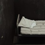 The Sleep-Deprivation Trap: Why 'Grind Culture' Is Sabotaging Your Competitive Edge Single bed with striped mattress against a dimly lit wall, creating a somber atmosphere.