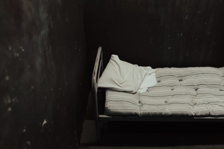Single bed with striped mattress against a dimly lit wall, creating a somber atmosphere.