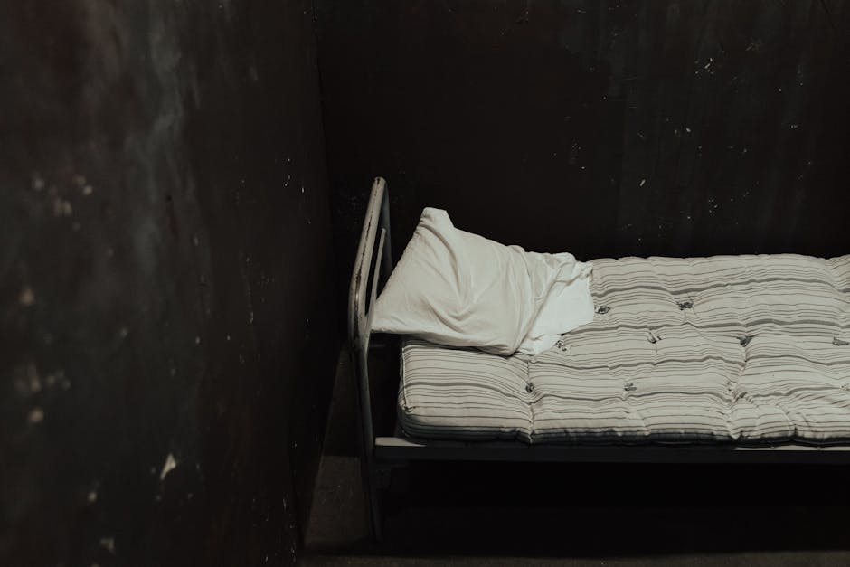 Single bed with striped mattress against a dimly lit wall, creating a somber atmosphere.