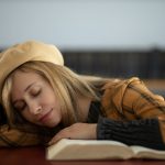 A young woman in a library naps on an open book with eyes closed, wearing a beret hat.