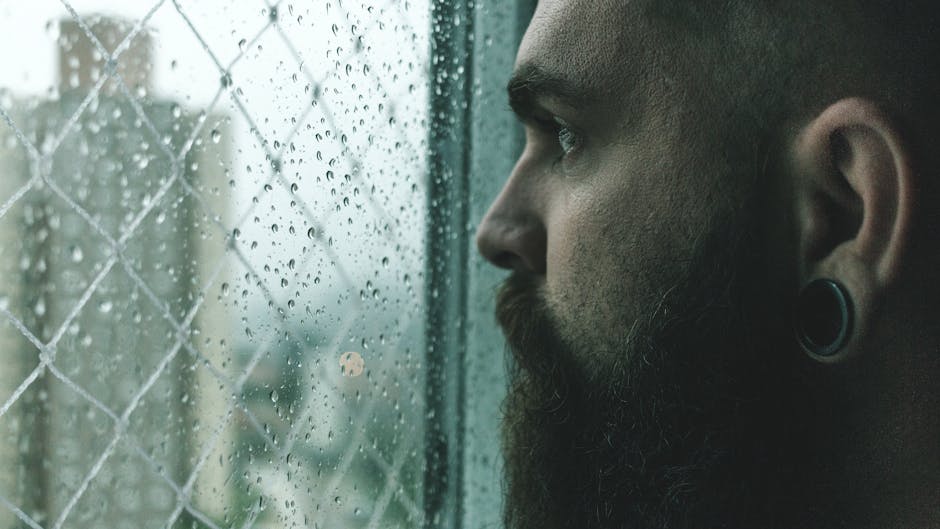 Close-up of a man with a beard looking thoughtfully through a rain-spattered window.