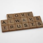 Close-up of wooden Scrabble tiles spelling 'Love Always Trusts' on white surface.