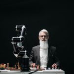 A bearded man in suit playing chess with robotic arm, showcasing AI strategy.