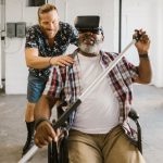 Virtual Reality Therapy: Operationalizing Psychological Resilience Man assists wheelchair user engaged in rehabilitation exercise with VR set indoors.