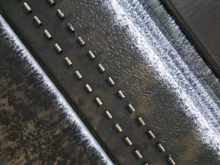 Aerial view of a structured water flow over a weir with rhythmic patterns and concrete blocks.