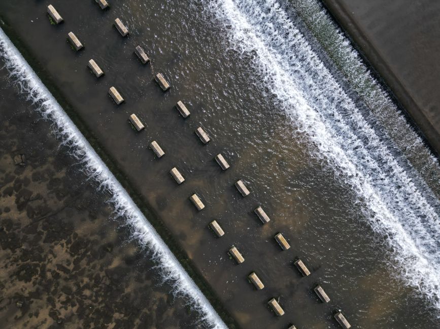 Aerial view of a structured water flow over a weir with rhythmic patterns and concrete blocks.