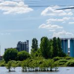 Scenic industrial landscape featuring a wind turbine, factories, and lush greenery by a body of water.