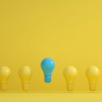 A standout blue light bulb among several yellow bulbs on a yellow background, symbolizing innovation.